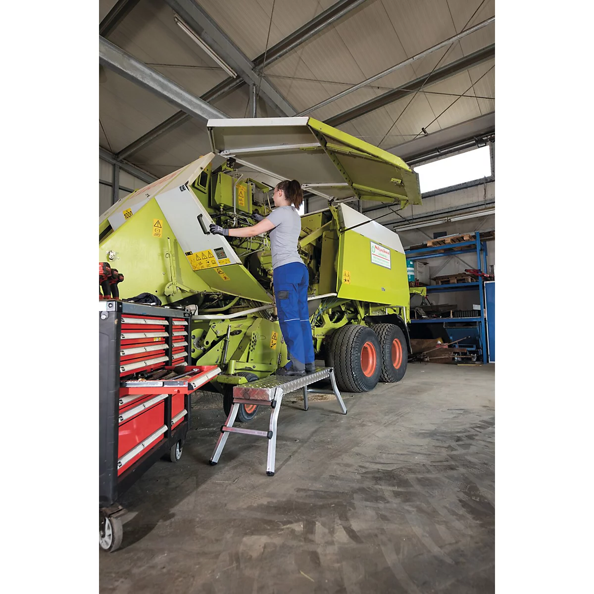 Een vrouw repareert een groene landbouwmachine in een werkplaats. Ze staat op een opstapje en opent een klep.