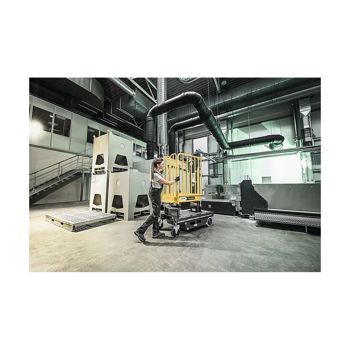 Une femme tire un chariot jaune dans un hall de production. Machines grises et tuyaux en arrière-plan.