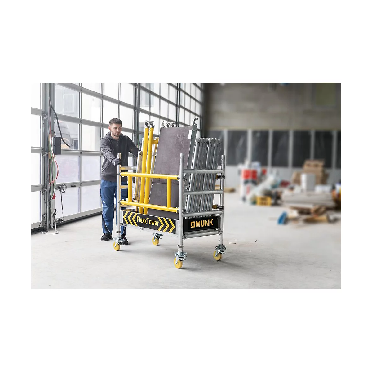 Een man duwt een transportkar voor een steiger. De kar bevat steigermateriaal, met het opschrift FlexxTower en Munk.
