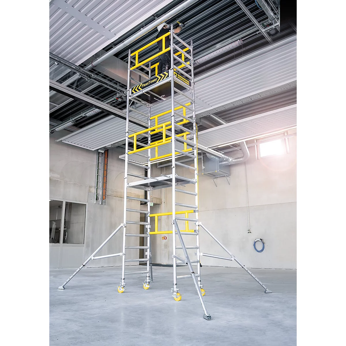 Een steiger met gele veiligheidsleuningen staat in een industrieel gebouw. Bovenin is een persoon te zien.