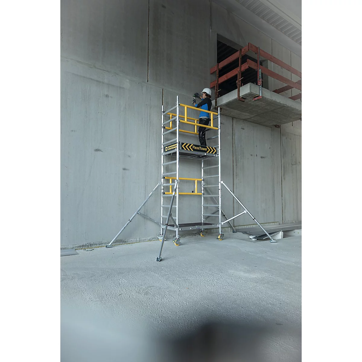 Persoon op een steiger voor een betonnen muur. Gele en zwarte markeringen op de steiger. Rode balk op de achtergrond.