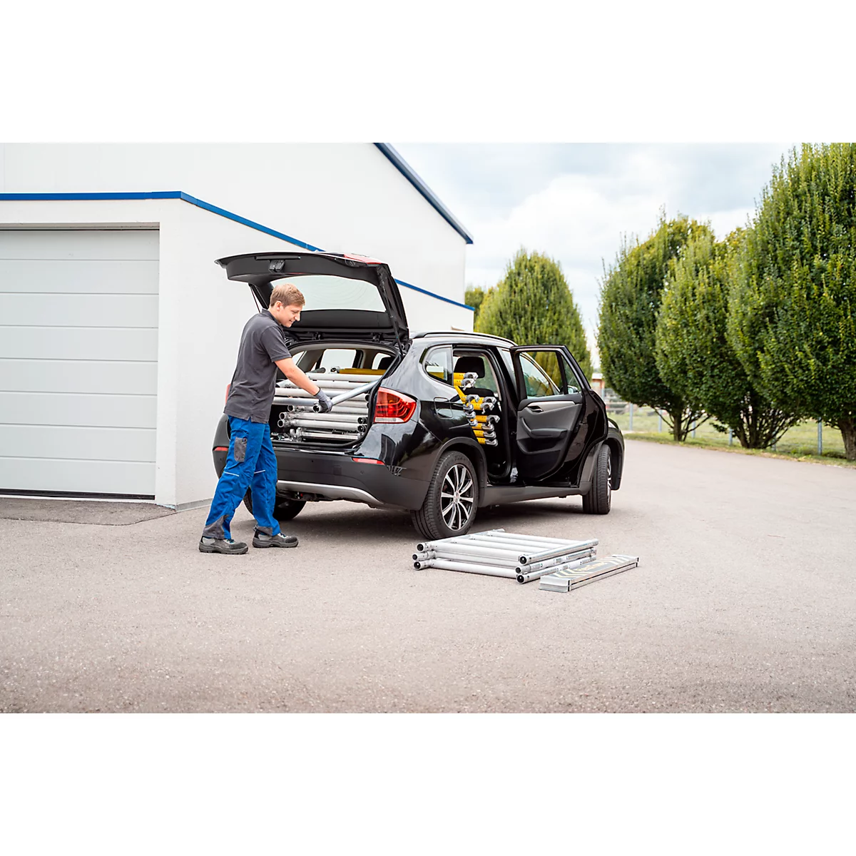 Een man laadt een ladder uit een zwarte auto. Andere ladderonderdelen liggen op de grond.