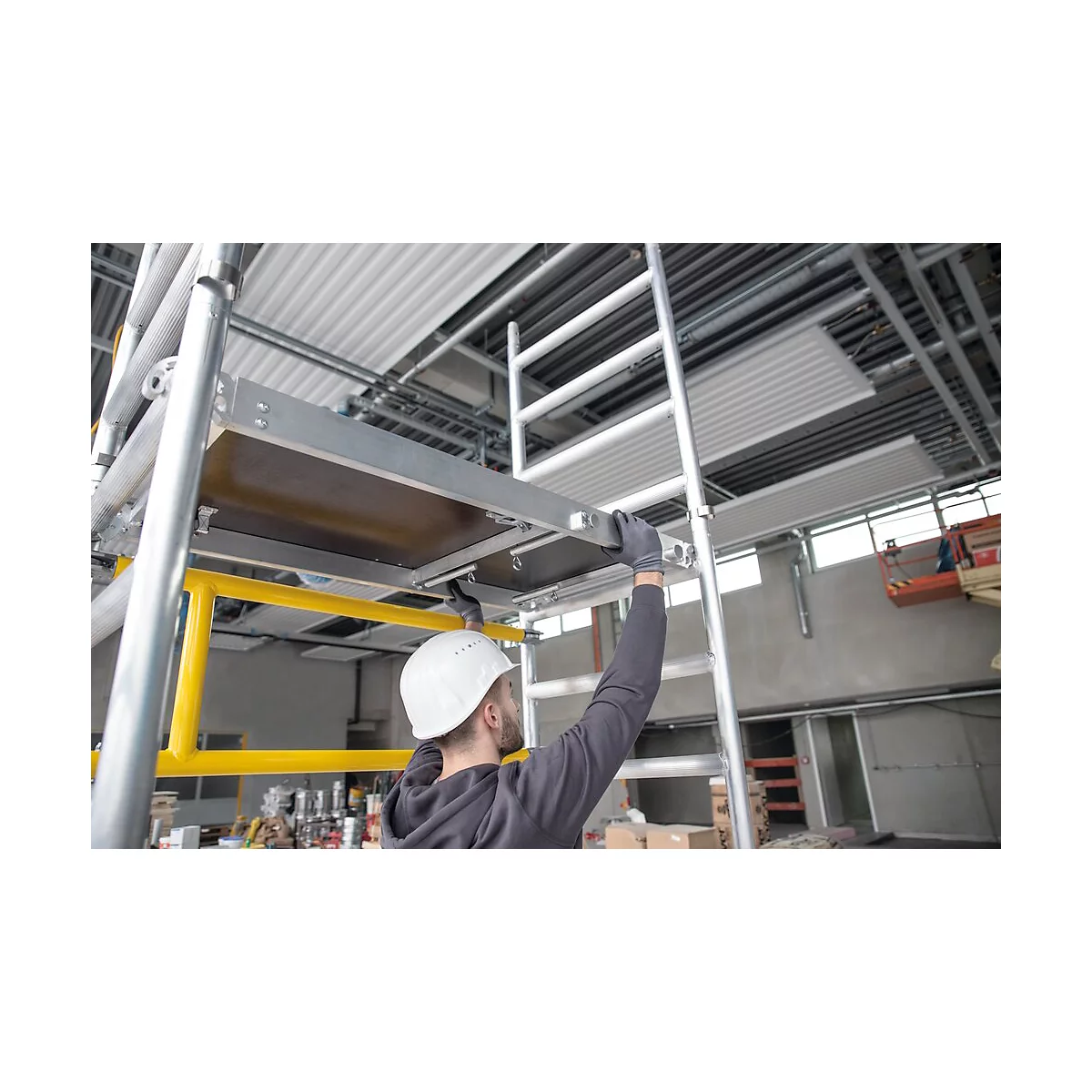 Een man met helm en handschoenen monteert een steiger. Hij staat op een ladder en heft zijn hand naar een platform.