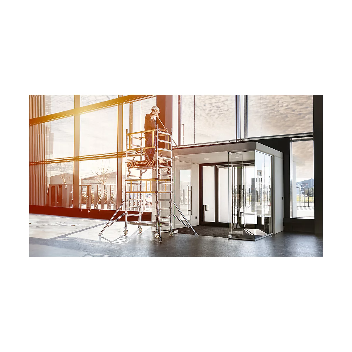 A man on scaffolding in a building is installing something on a glass door. Sunlight streams through the windows.