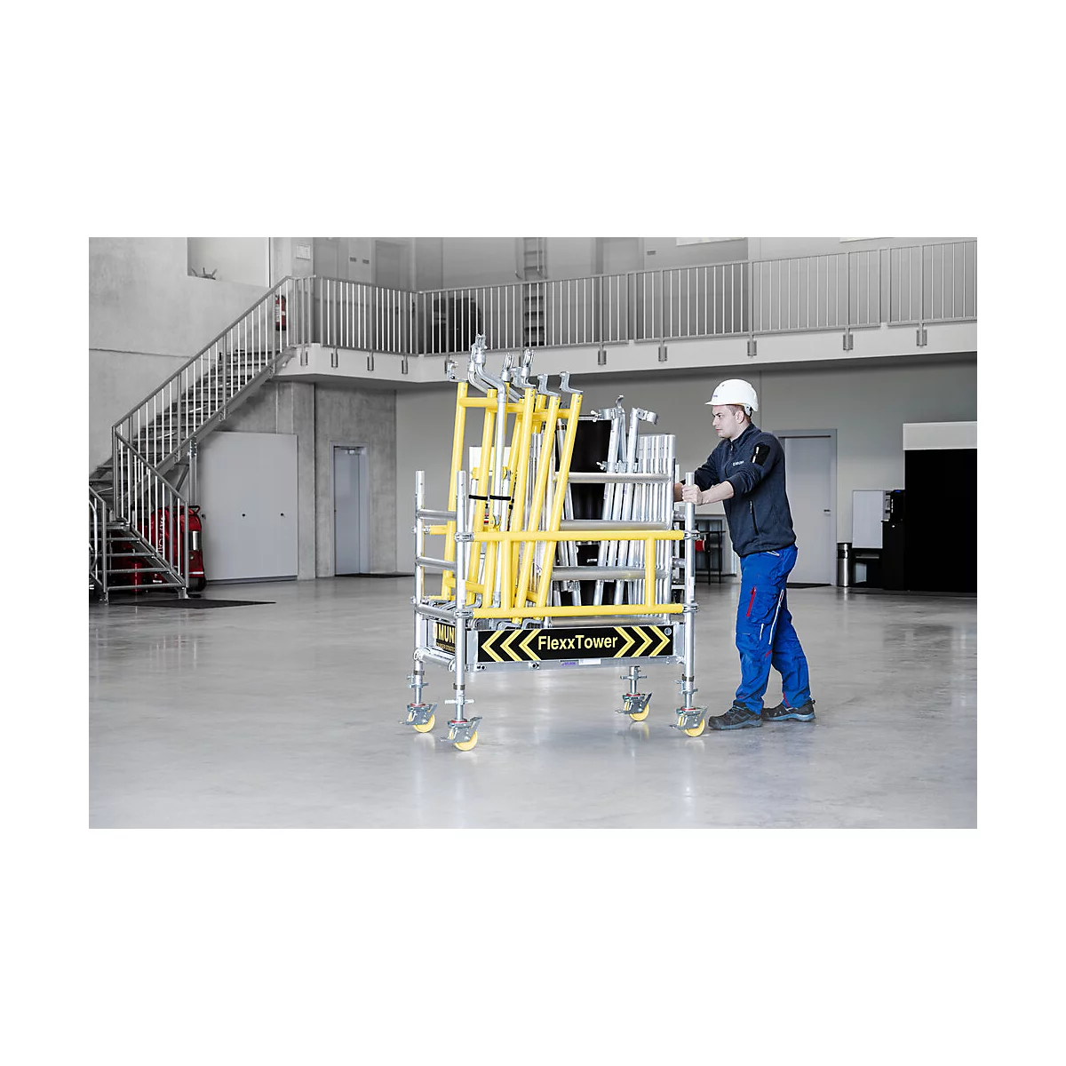 A man pushing a yellow FlexxTower scaffolding cart on wheels in a warehouse. A staircase in the background.