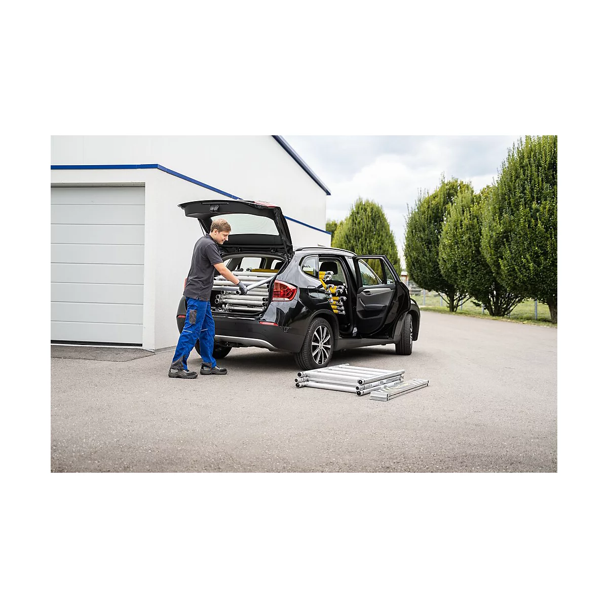 A man loads ladder parts into a black station wagon. More ladder parts are lying on the asphalt.
