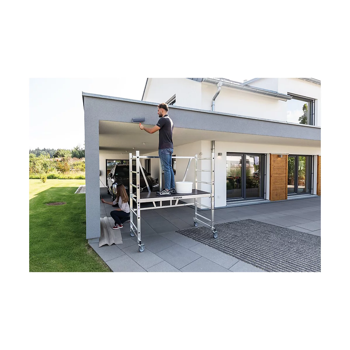 Man paints the outside wall with a roller, woman paints on the side. Both are standing on a scaffolding platform.