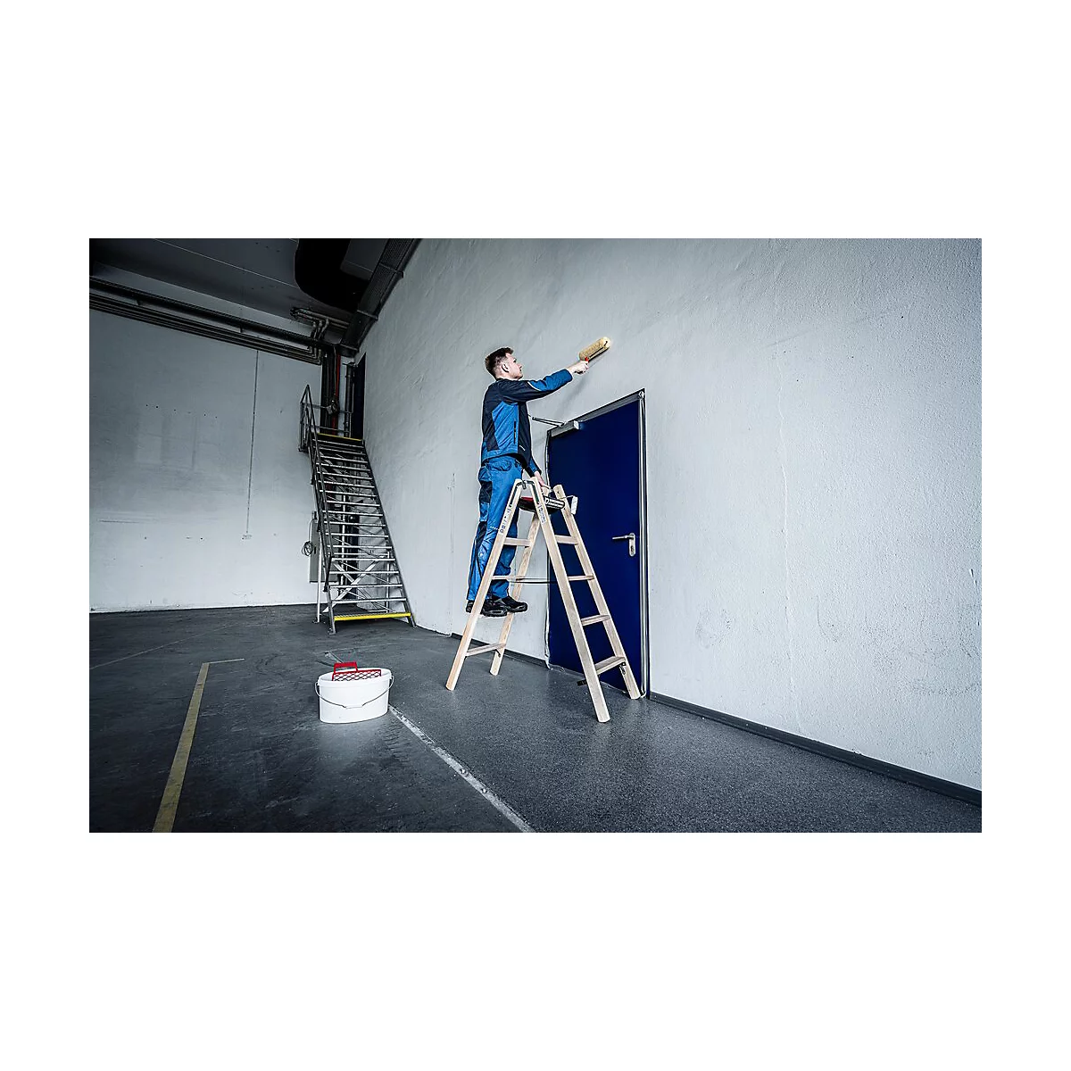 Ein Mann in blauer Arbeitskleidung streicht eine Wand. Er steht auf einer Leiter, hinter ihm eine blaue Tür.
