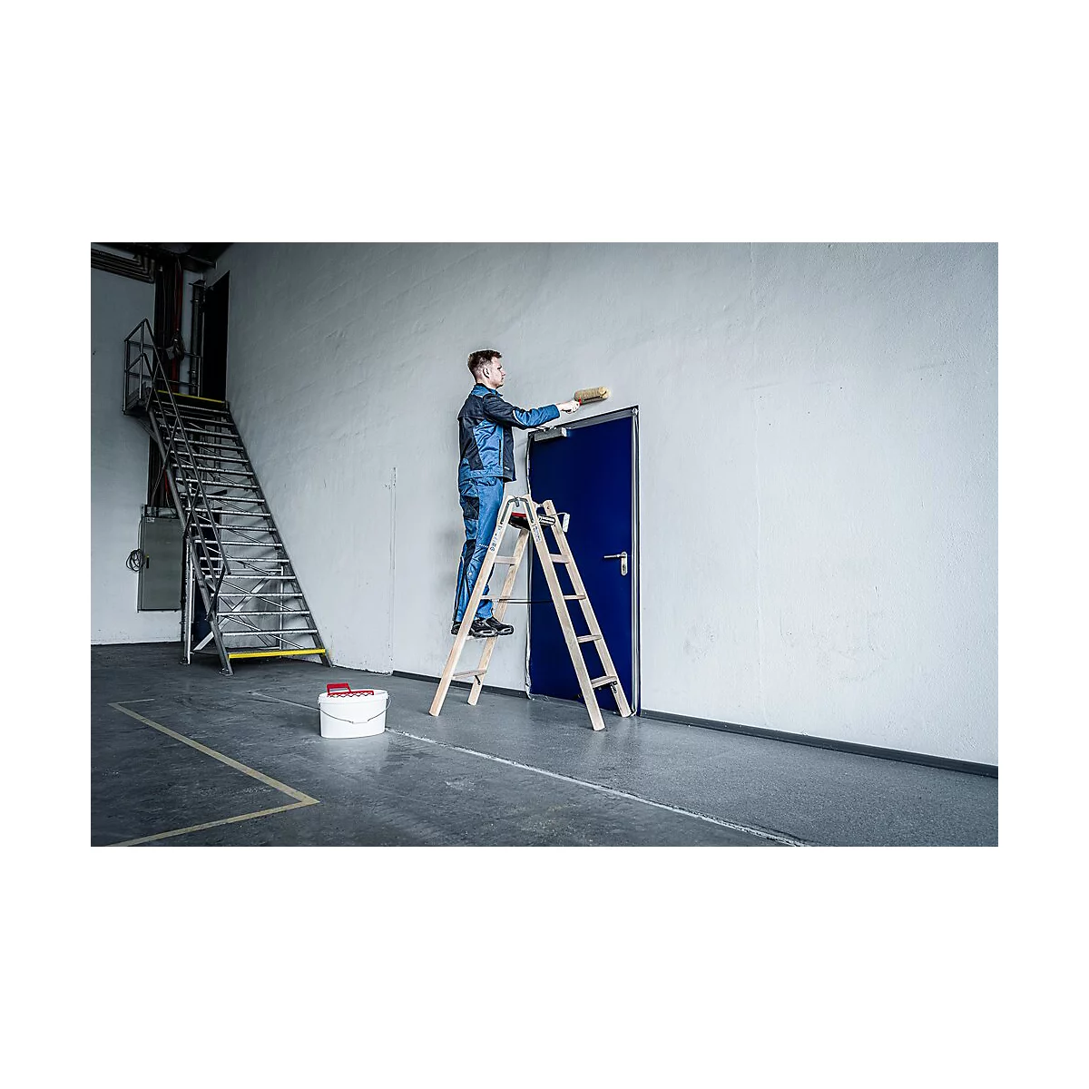 Een man schildert de muur boven een blauwe deur, staand op een ladder. Rechts een witte muur, links een trap, een emmer en gereedschap op de grond.