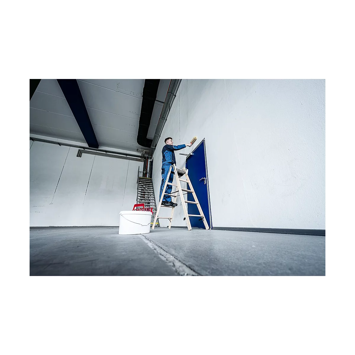 Man op een ladder schildert een blauwe deur en witte muur. Emmer en verf staan ernaast.
