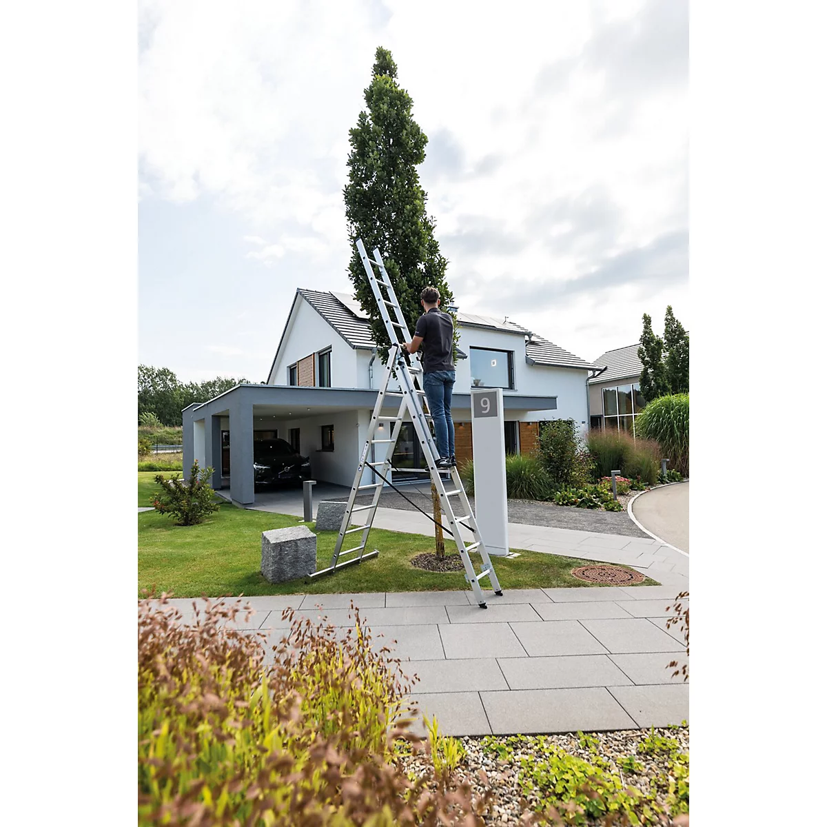 Man op een ladder die een boom snoeit. Achter hem: huis met carport, gazon, verharde oprit.