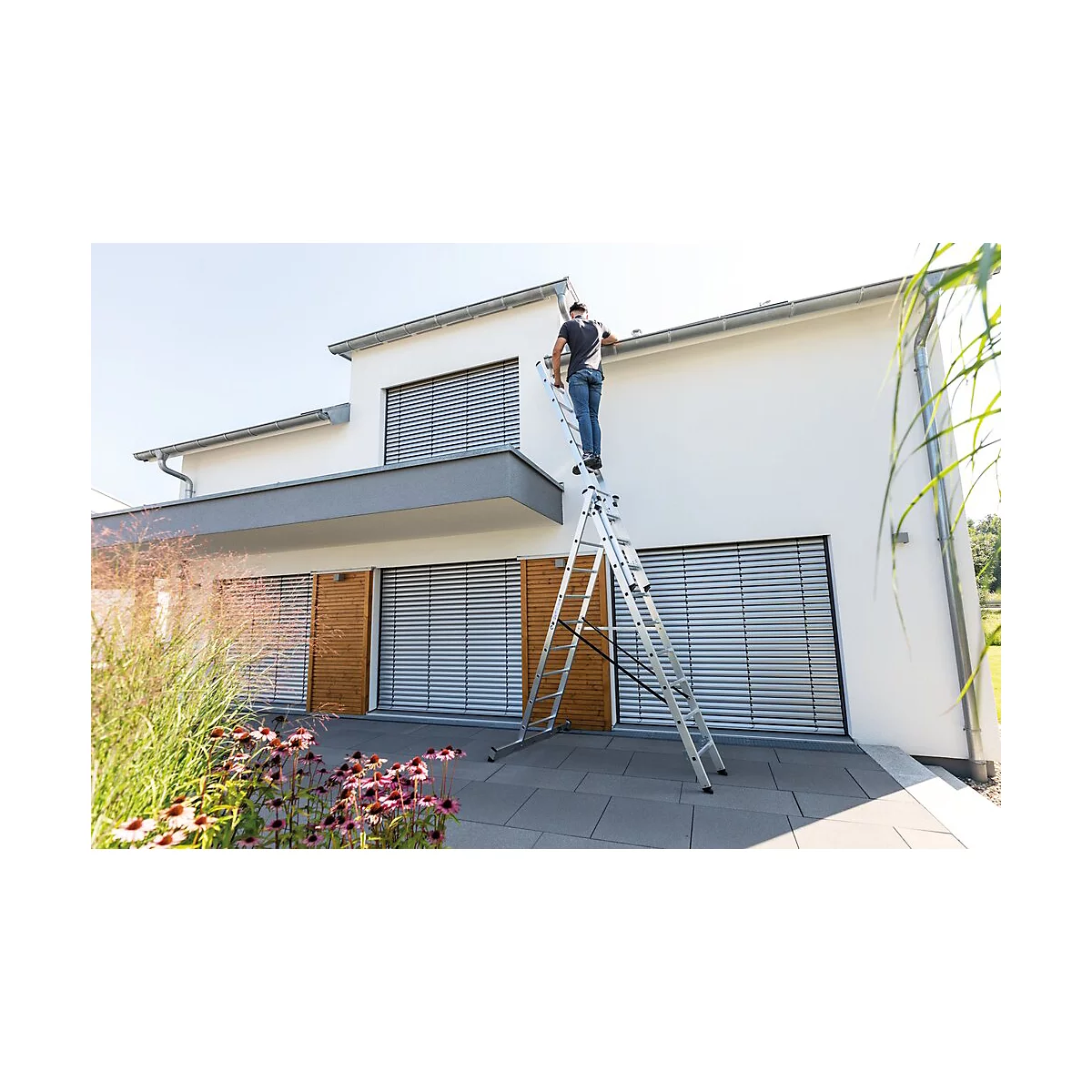 Man op ladder bij wit huis. Hij werkt aan de dakgoot. Bloemen en struiken vooraan.