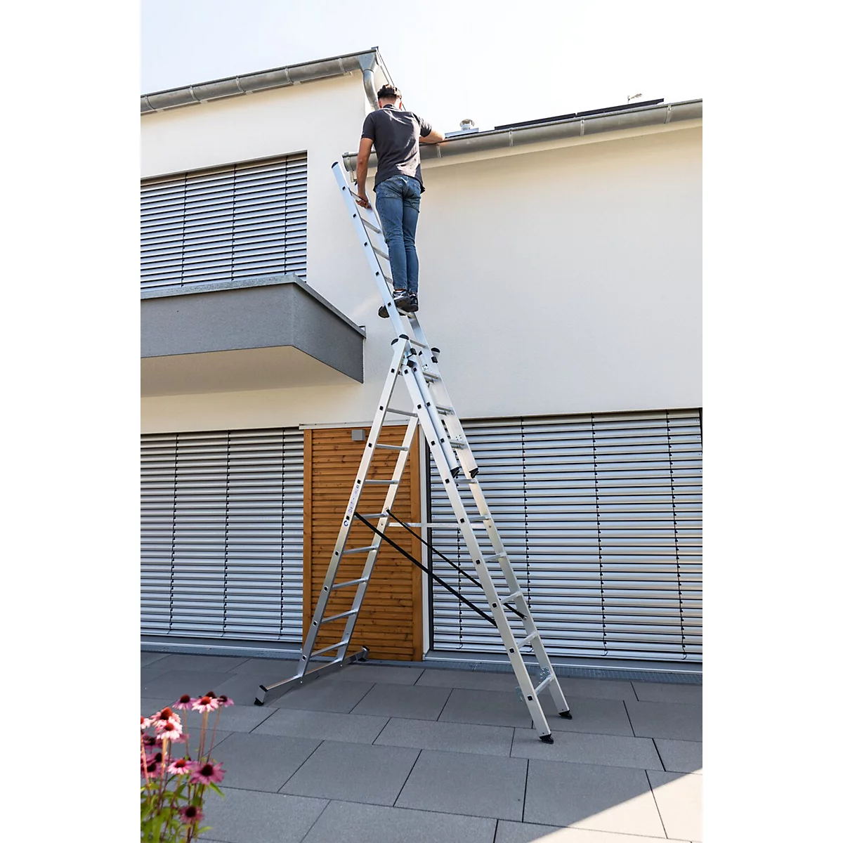 Man staat op een ladder bij een huis. Hij werkt aan de dakgoot. Bloemen op de voorgrond, gevel van het huis, ramen.