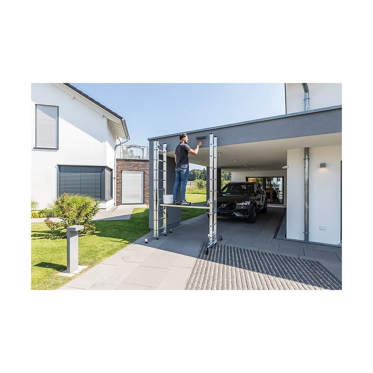 Man schildert de muur van het huis, staand op een steiger. Op de achtergrond een garage met een auto en een huis.