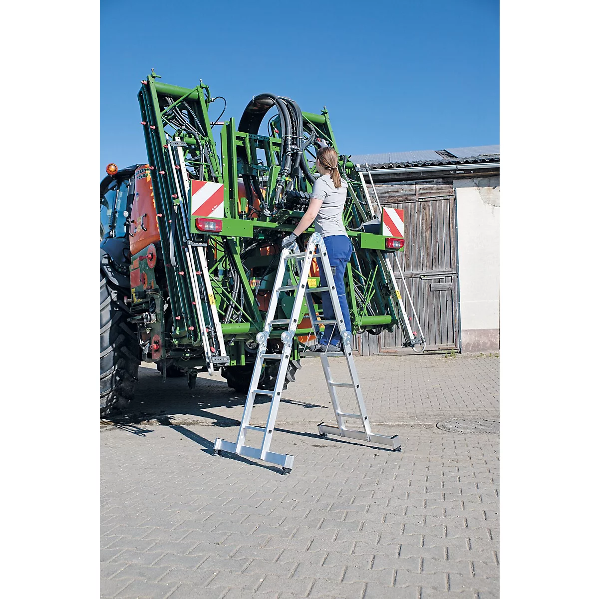 Een vrouw beklimt een ladder op een tractor. De tractor heeft een groene sproeier, de lucht is blauw.