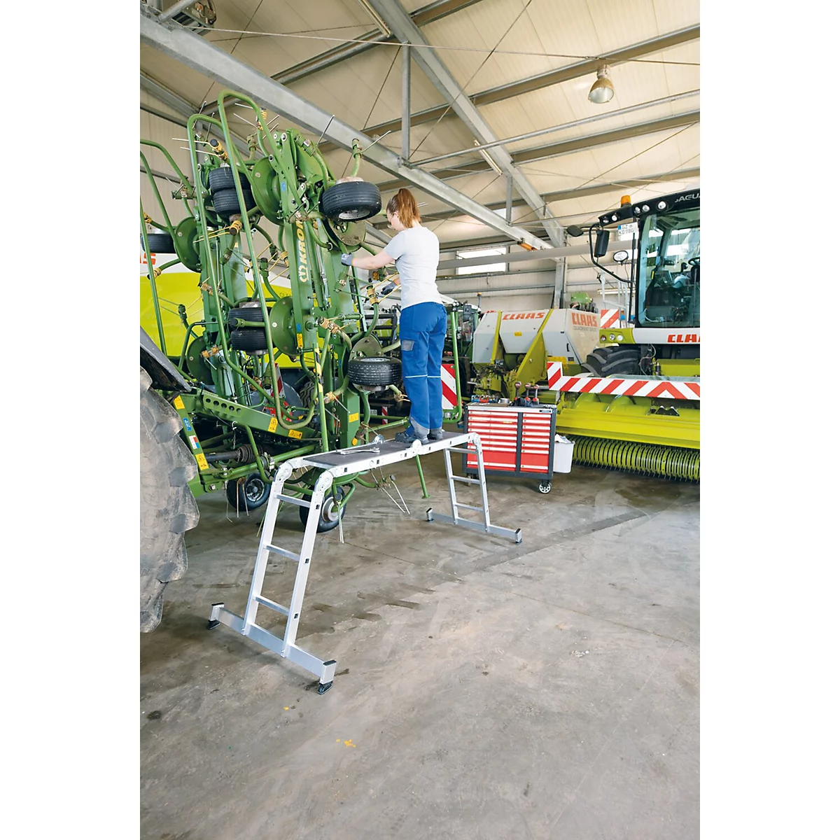 Een persoon op een trap werkt aan een tractor in een werkplaats. Andere machines op de achtergrond.