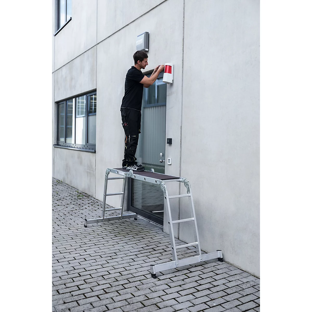 Een man werkt aan een muur, staand op een werkplatform. De muur is wit en heeft een deur en een raam.