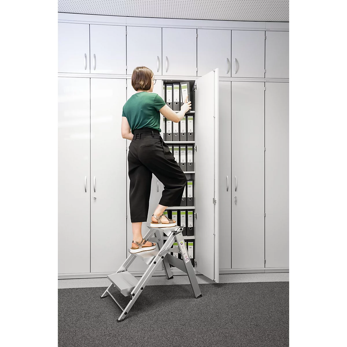 Een vrouw staat op een trap en reikt in een open archiefkastcompartiment. Grijze kasten op de achtergrond.