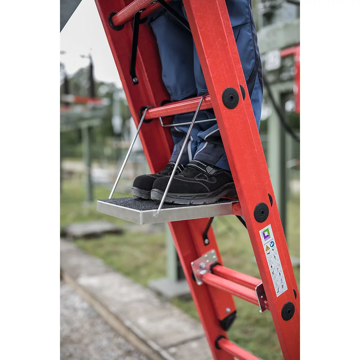 Detailopname van een rode ladder met voeten in werkschoenen erop. Duidelijk zichtbaar loopvlak.