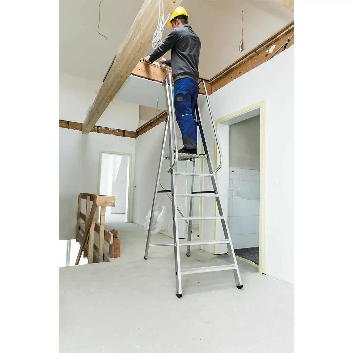 Een man op een ladder werkt aan een plafondconstructie. Hij draagt een gele helm en werkkleding.
