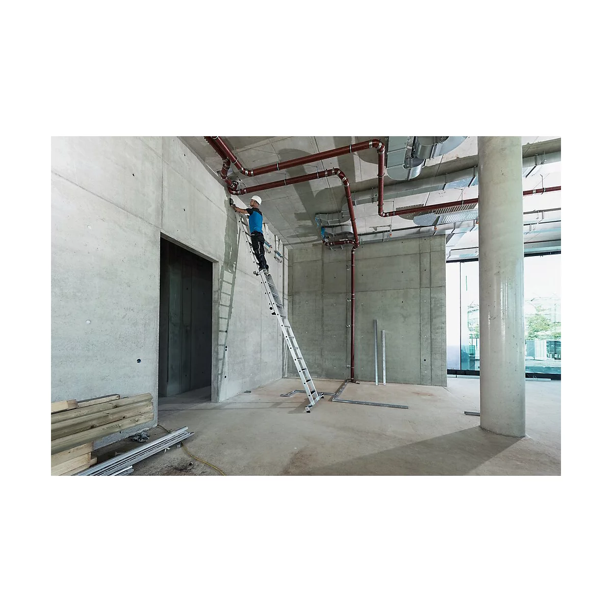 Ein Handwerker steht auf einer Leiter und arbeitet an Rohren, die an der Decke eines Rohbaus befestigt sind.