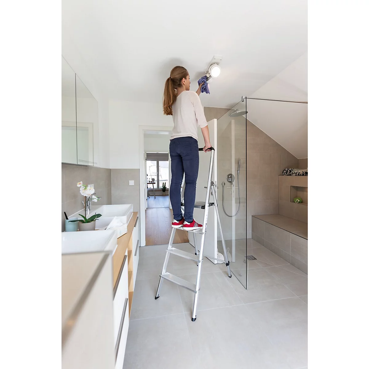 Frau putzt im Bad die Deckenlampe mit einem Tuch. Sie steht auf einer Trittleiter.