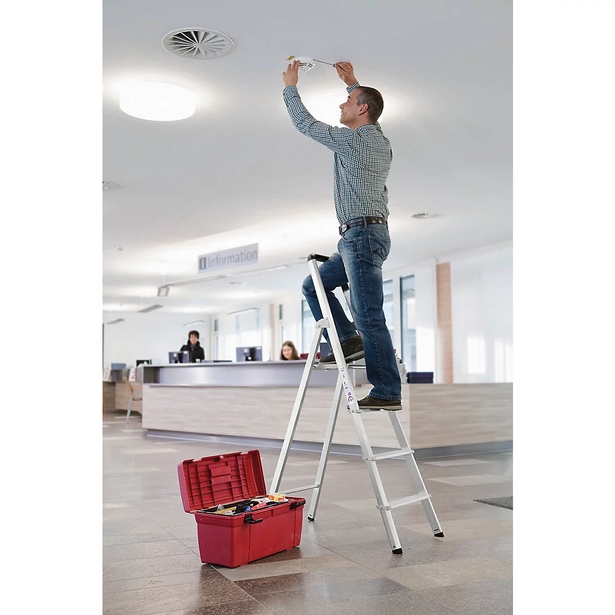 Ein Handwerker auf einer Leiter repariert eine Deckenleuchte. Roter Werkzeugkasten am Boden, im Hintergrund eine Rezeption.