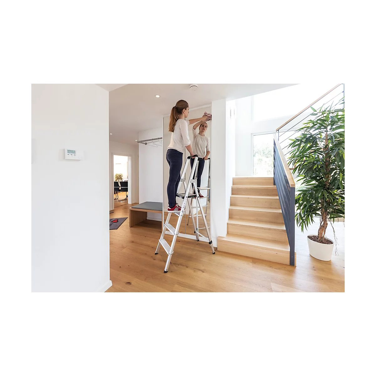 Frau steht auf einer Trittleiter, um ein Bild aufzuhängen. Spiegel zeigt Frau, Holzfußboden und Treppe.