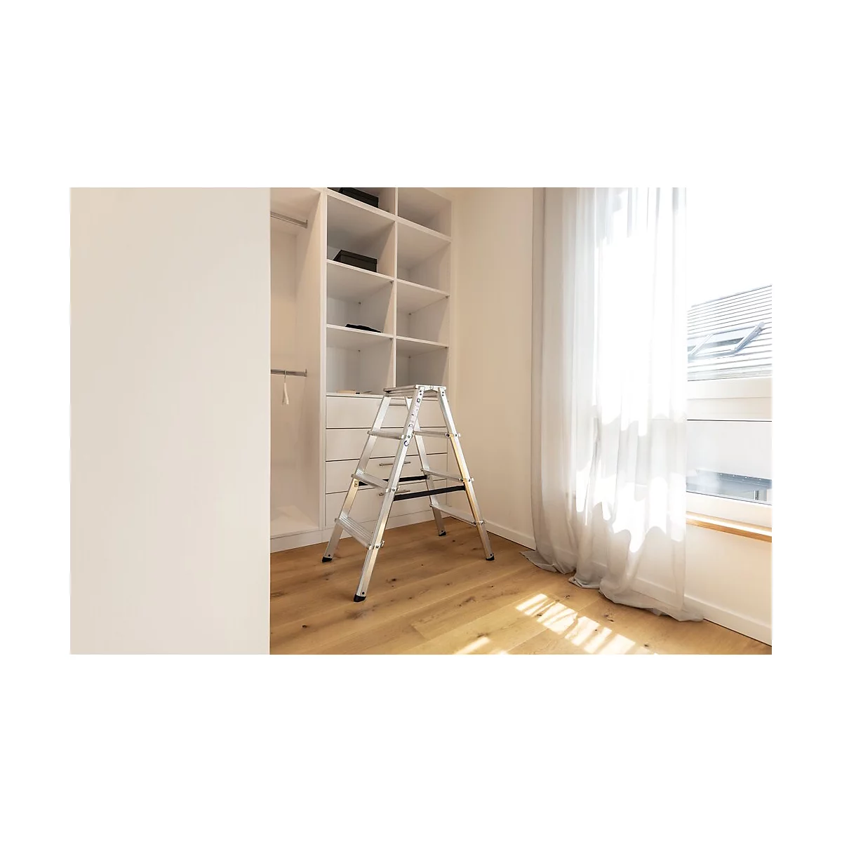 Ein silberfarbener Trittschemel steht vor einem weißen Einbauschrank. Rechts ein Fenster mit Vorhängen.