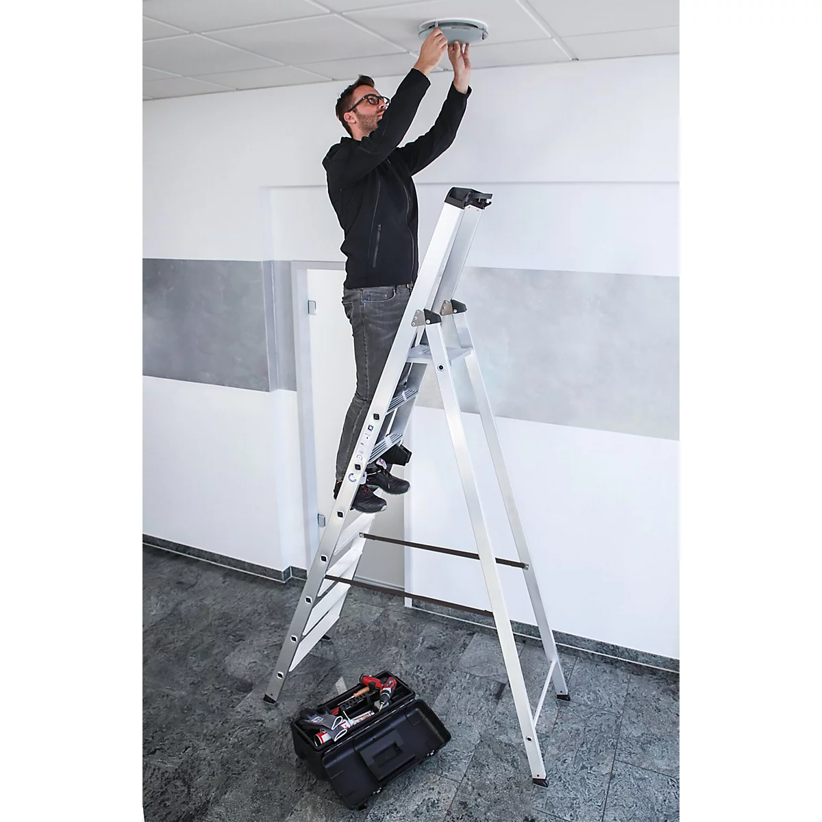 Man op een ladder die een plafondlamp monteert. Gereedschapskist op de vloer.