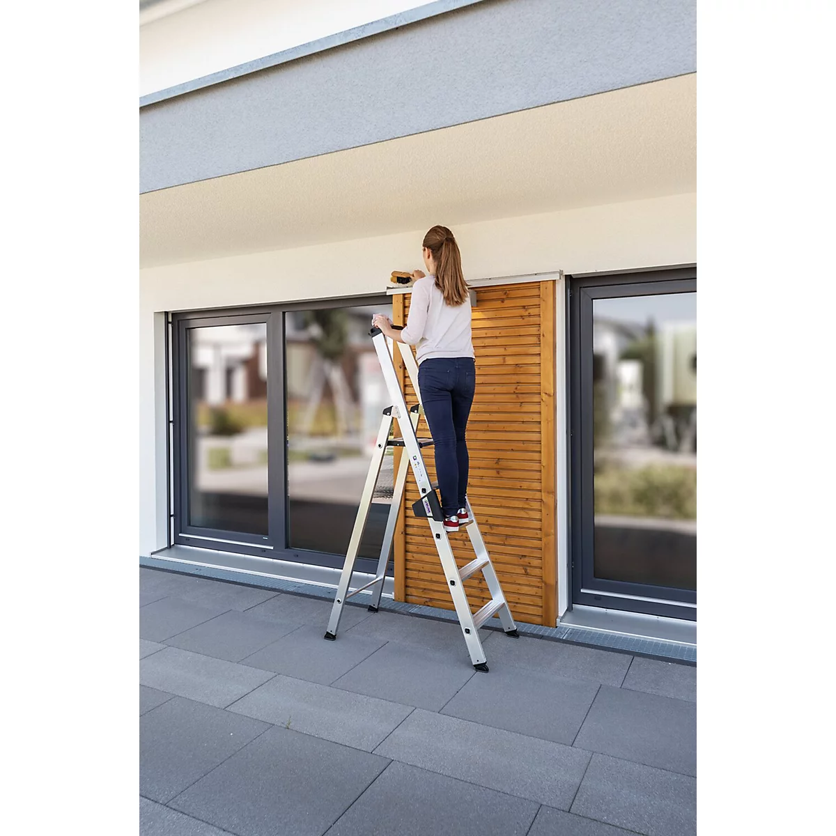 Een vrouw staat op een ladder en werkt buiten aan een houten bekleding.