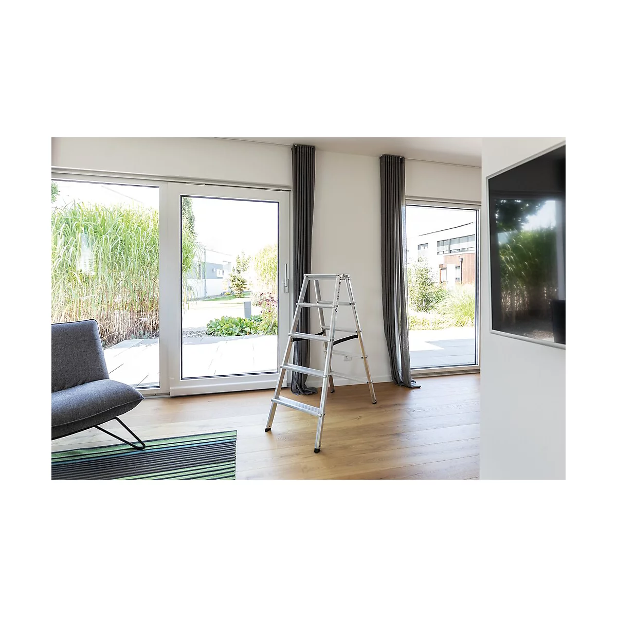 Een kleine ladder staat in een moderne woonkamer. Grote ramen bieden uitzicht op de tuin. Een televisie hangt aan de muur.