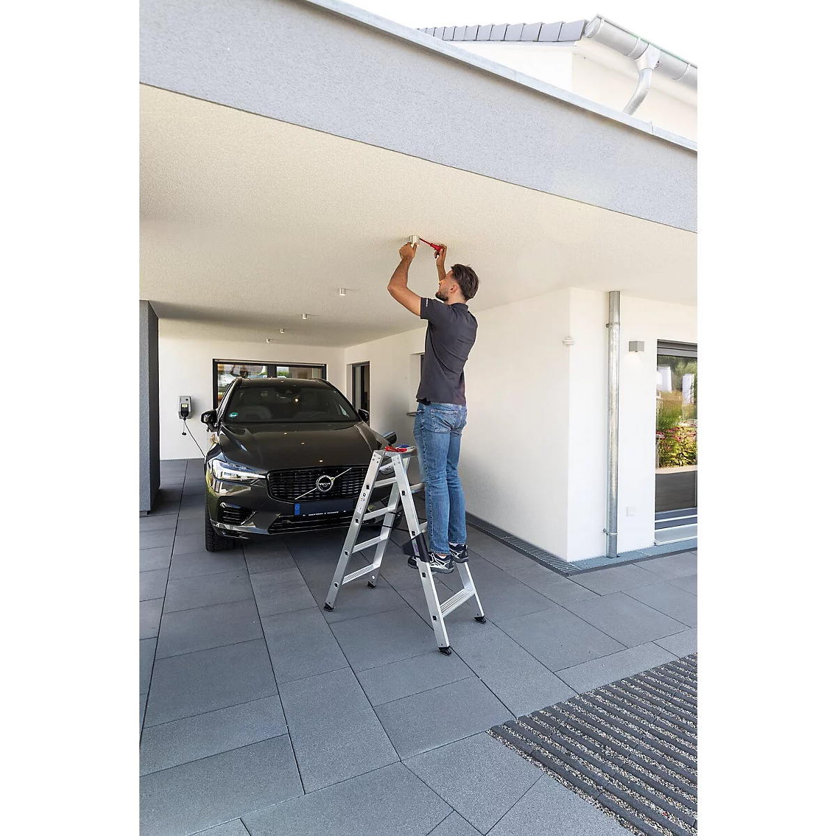 Een man op een ladder installeert een plafondlamp aan het plafond van een garage met een zwarte auto erin.