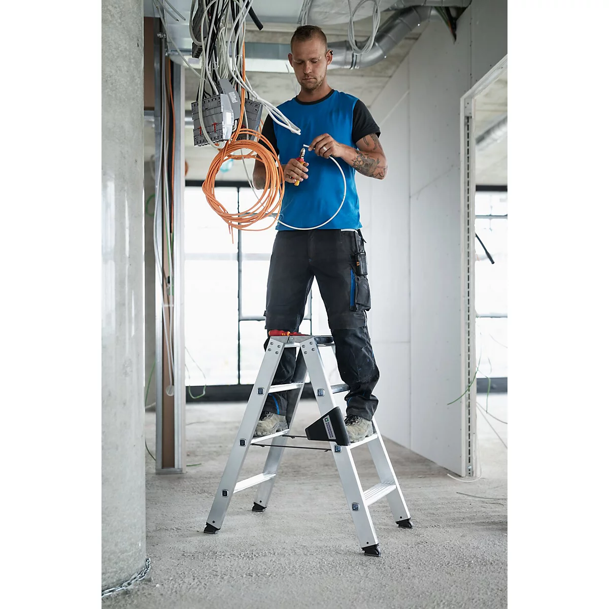 Elektricien op een trap, kabels aan het installeren in een casco gebouw. Hij draagt werkkleding, kabels en plafond op de achtergrond.