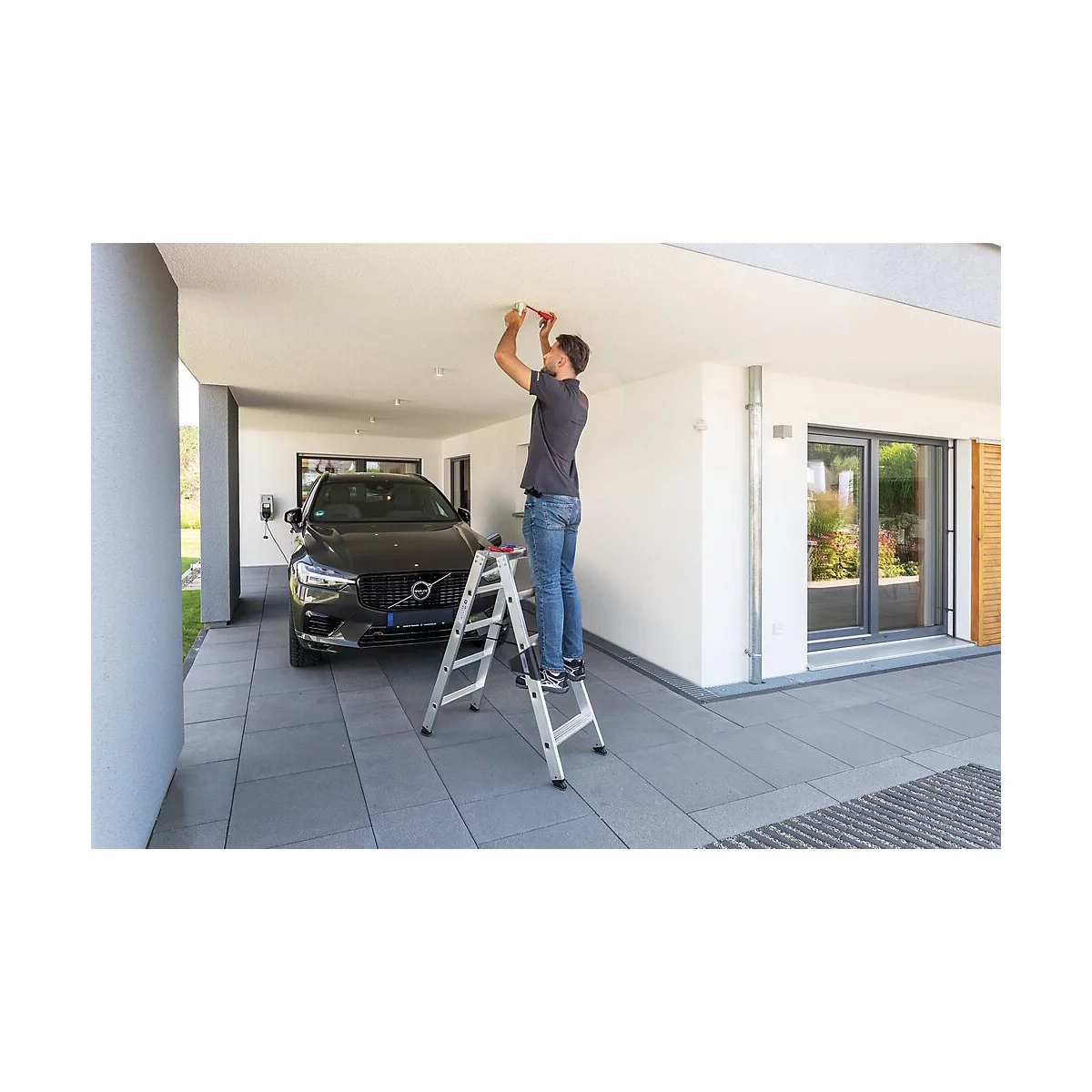Man op een ladder die licht in de carport installeert; een zwarte auto geparkeerd achter.