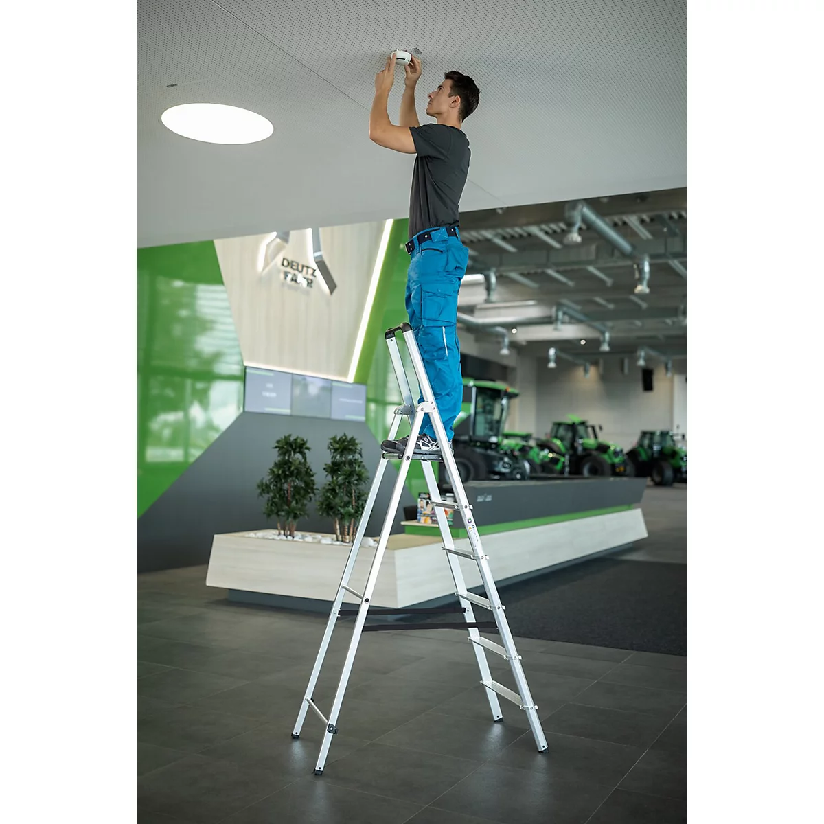 Een man op een ladder installeert een lamp aan het plafond. Op de achtergrond: groene tractoren en het Deutz-logo.