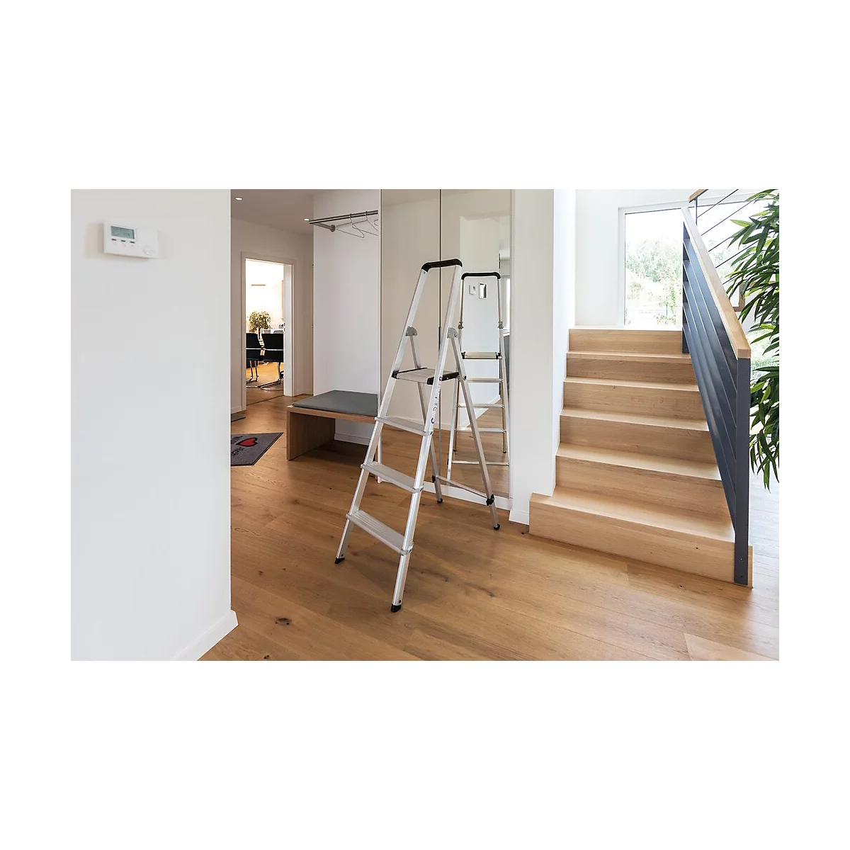Een zilveren trap staat in een hal. Rechts een houten trap, links een bank en een spiegel.
