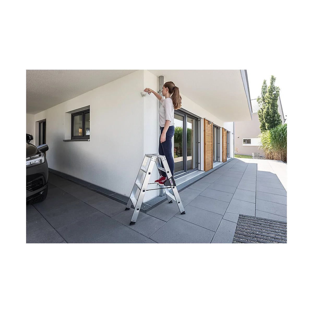 Vrouw op een ladder die een camera aan de muur van het huis bevestigt. Wit huis met grijze grond. De blik van de vrouw is gericht op de camera.