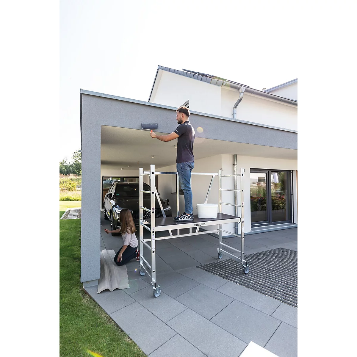 Een man schildert het plafond van een garage, terwijl een meisje op de grond zit. Een ladder staat klaar.