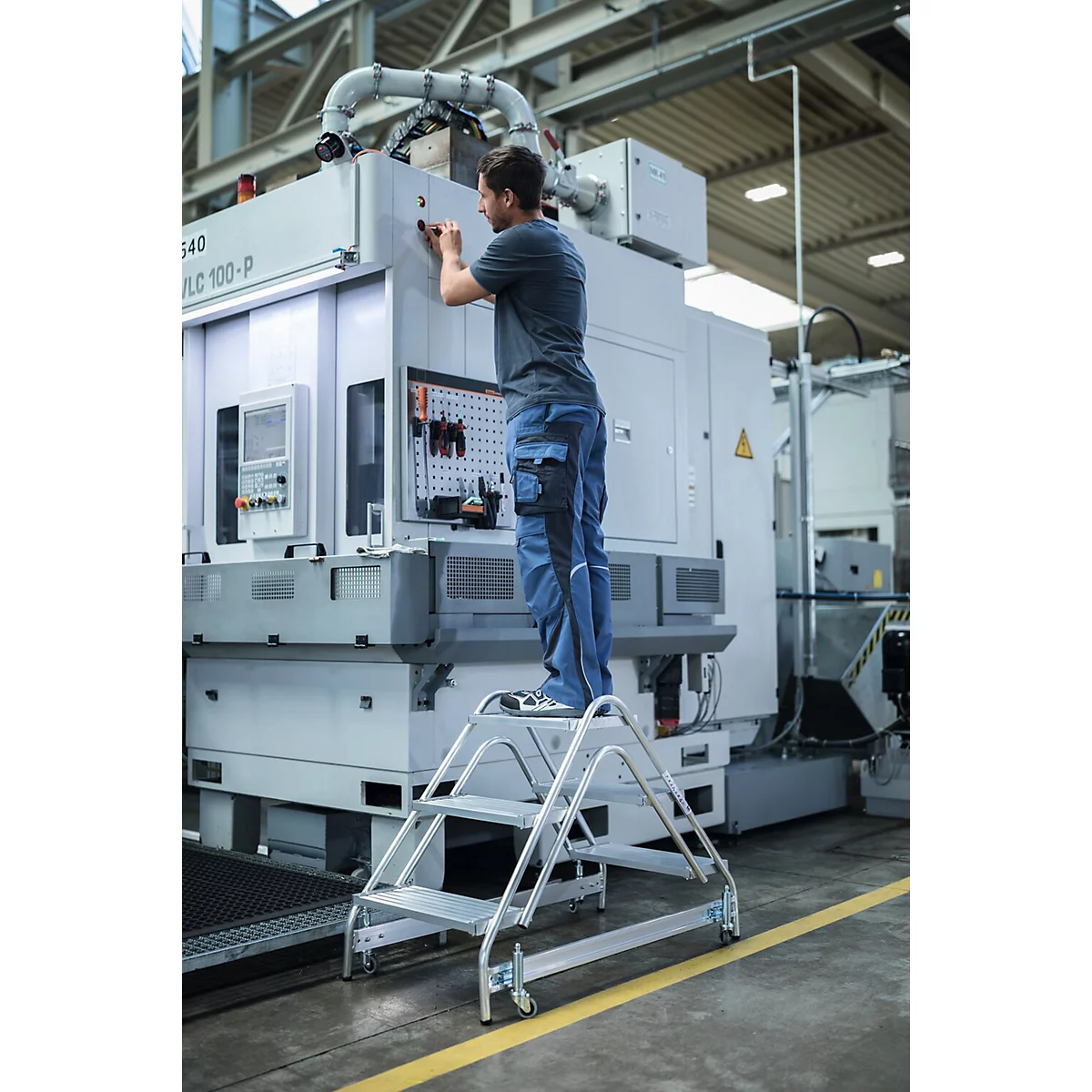Man op een trap monteert machine in een fabriek. Grijze trap, blauwe broek.