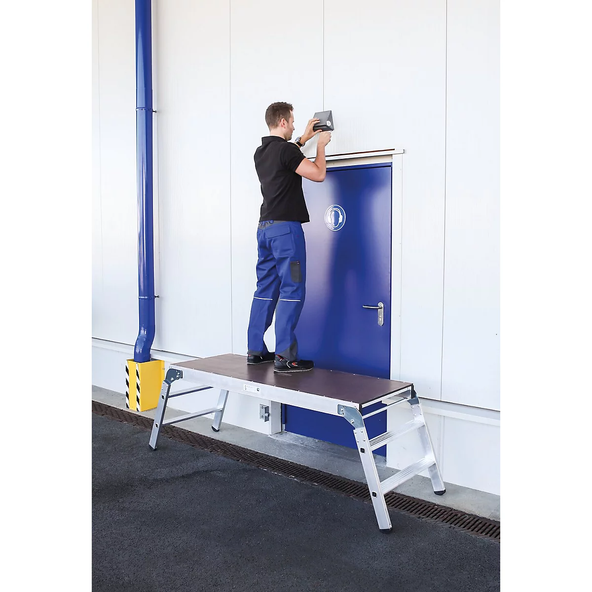 Een vakman op een werkplatform monteert iets aan een blauwe deur. De scène speelt zich af in de openlucht.