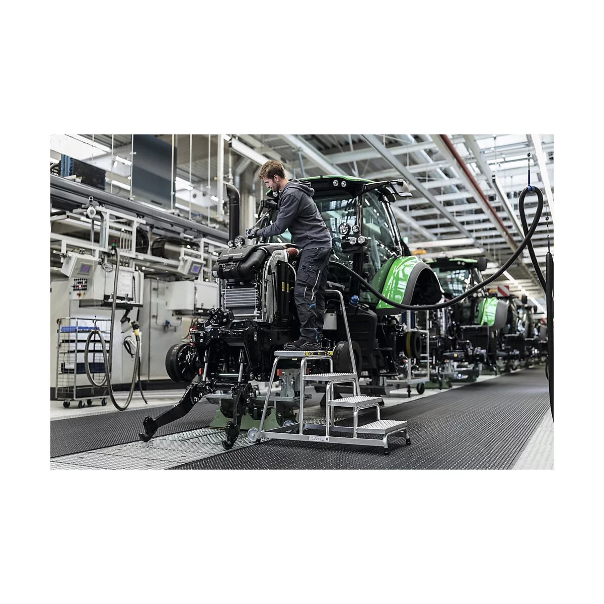 Een man werkt aan een tractor in een fabriek, staand op een kleine trap.