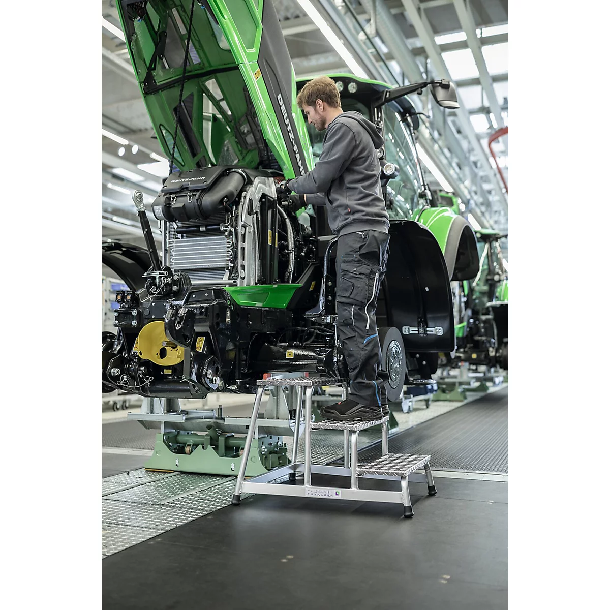 Een man werkt aan een groene tractor, staand op een trap. In een fabriekshal, open motorkap.