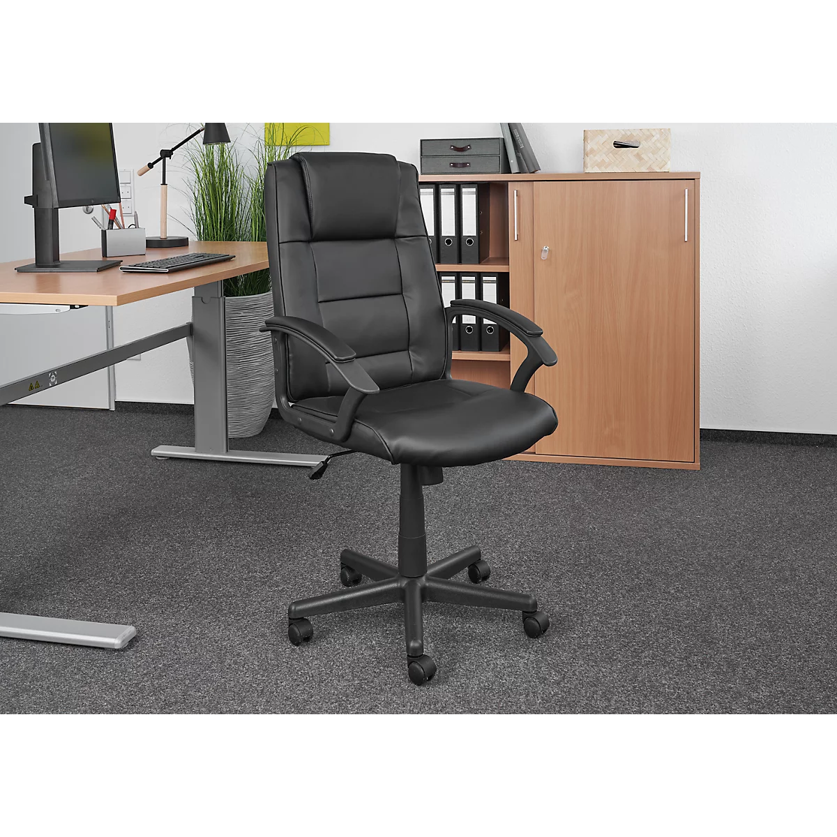 Black office chair in front of a desk and cabinet. The chair is centered, with a padded back, armrests, and casters.