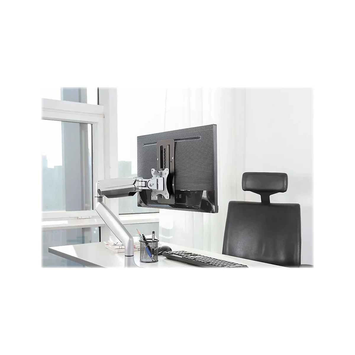 Ein Monitor ist an einer Halterung an einem Schreibtisch befestigt. Im Hintergrund ist ein Stuhl und ein Fenster zu sehen.