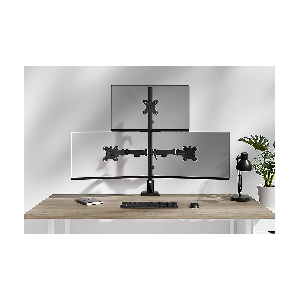 Drievoudige monitorstandaard met drie schermen op een bureau, naast plant en lamp, toetsenbord en muis.