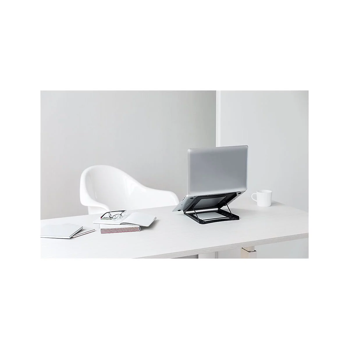 Ein Laptop auf einem schwarzen StÃ¤nder steht auf einem weiÃŸen Schreibtisch. Daneben ein weiÃŸer Stuhl, ein Buch und eine Tasse.