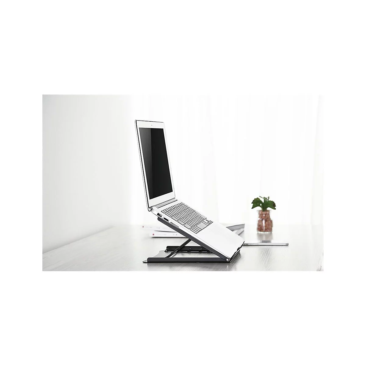 Ein silberner Laptop steht auf einem schwarzen Laptop-StÃ¤nder, der auf einem Tisch platziert ist. Im Hintergrund eine kleine Pflanze.
