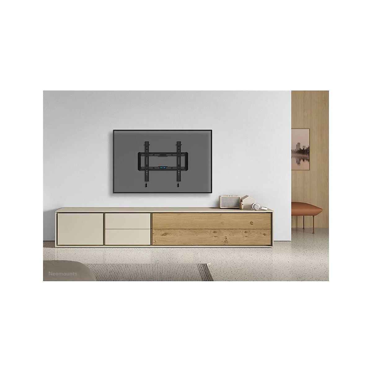 Ein TV-Wandhalter, montiert an einer grauen Wand, über einer modernen Kommode, mit Dekor. Die Marke Neomounts ist unten links zu sehen.