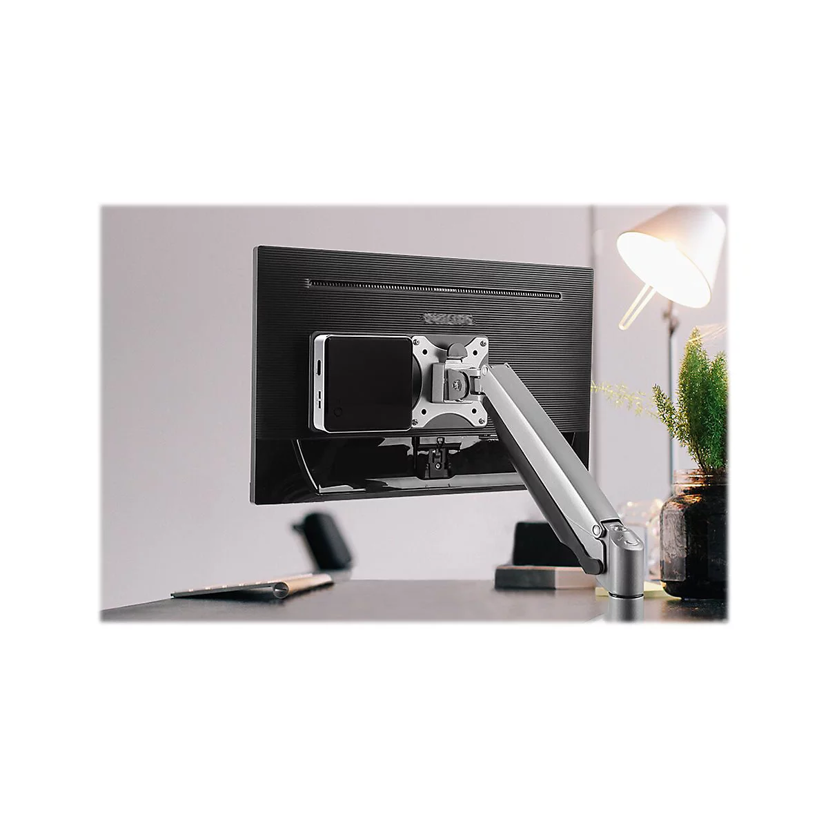 Ein Monitor mit Halterung und darauf befestigtem GerÃ¤t. Im Hintergrund ein Schreibtisch mit Lampe und Pflanze.